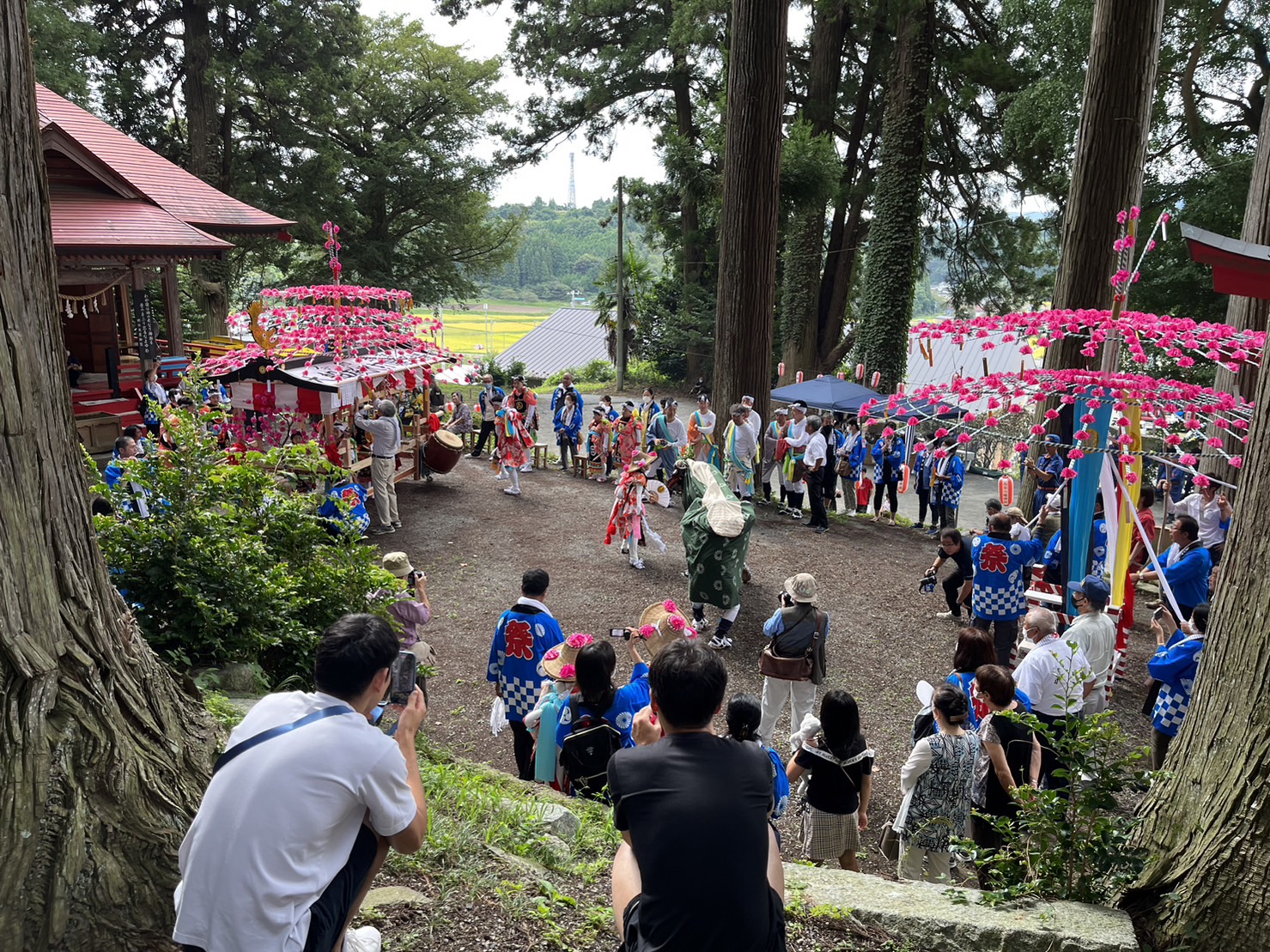 山間の神社の境内で、赤や青のはっぴを着た人々が太鼓を叩いたり舞を踊ったりしており、たくさんの観客が木々の間から見守っている祭りの写真