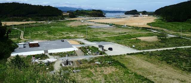 中央には石像や石灯篭があり、周囲には草が生い茂る平地が広がり、遠くに港と海が見える高台から写したパノラマ写真