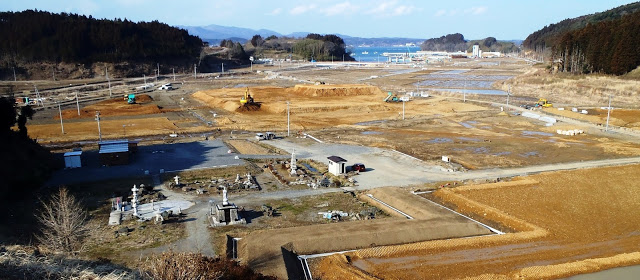 中央には石像や石灯篭があり、重機が動き、茶色い土がむき出しになった広大な土地で、道路や区画整理が進められている復旧工事のパノラマ写真