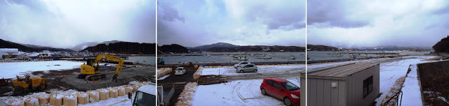 雪が積もった岸辺に重機や車が停まり、正面の海には漁船が並ぶ冬の漁港のパノラマ写真