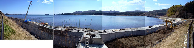 青空のもと海が広がりコンクリート製の防潮堤に鉄筋が並んでクレーン車で工事中のパノラマ写真