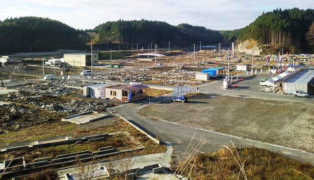 周囲を山々に囲まれた、建物の基礎や瓦礫が多く残り、仮設店舗や建物が点在している写真