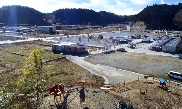 手前には子供向けの遊具が設置された小さな公園があり、その先には広い造成地が広がっている。造成地の中には仮設の建物が複数建てられており、その周囲にはたくさんのカラフルな旗が立てられている写真