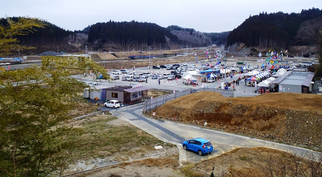 広場にはテントが立ち並びカラフルな旗が立ち、人々が集まり祭りの様子を写した写真