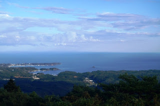 山の上から見下ろす視点で、入り組んだ海岸線や町並みが広がり、遠くに青い海と空がつながって見える写真
