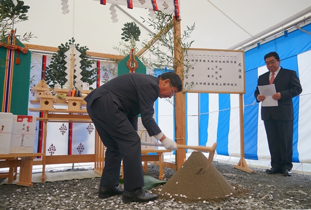 祭壇が設置され式次第が書かれた紙が掲示されている建設安全祈願祭で、スーツ姿の男性が鍬入れの儀を行っている写真