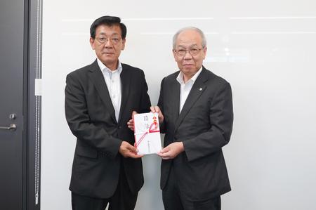 白い壁を背にしてスーツ姿の男性2人が並んで立ち、中央で熨斗のついた封筒を一緒に持ちながらカメラに向かって静かに微笑んでいる写真