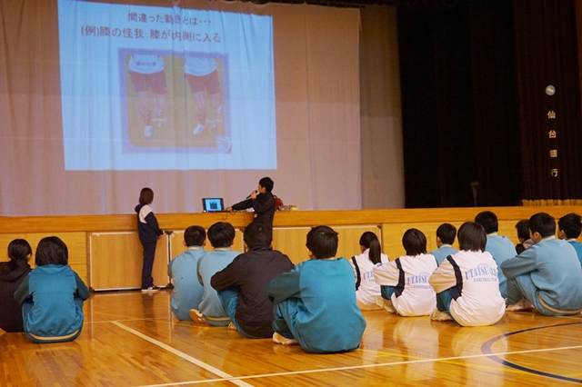 体育館のステージ前でジャージ姿の中学生たちが床に座ってスクリーンを見つめており、前方では理学療法士の講師の方がマイクを持ち、プロジェクターに映し出された正しい歩き方について説明をしている講話中の様子を写した写真