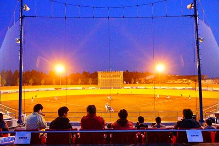 観客席から見た夜の野球場で、照明に照らされたグラウンド上で野球の試合が進行している様子を捉えた写真