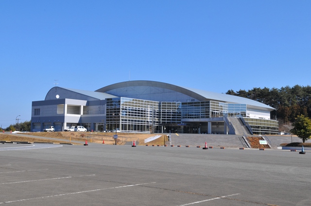 青空の下にある広い駐車場の奥に近代的なデザインで屋根が波のように湾曲した大きなガラス張りの南三陸町スポーツ交流村の外観写真