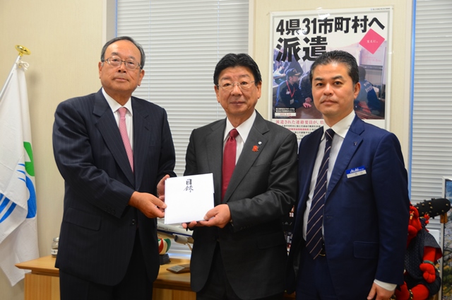 市長を中心に両隣にスーツを着た男性が立ち、左側の男性が市長に目録を手渡している写真