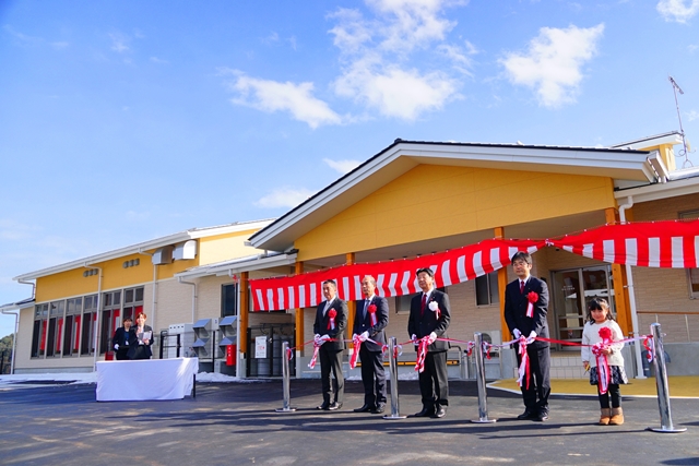 新しく建てられた建物の前で、赤と白のリボンを手にした4人のスーツ姿の男性と女の子が横一列に並び、テープカットを行おうとしている写真