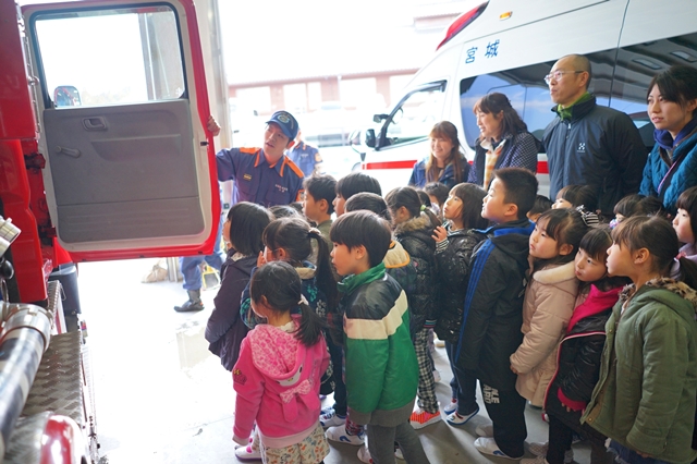 消防士が消防車のドアを開けて中を見せながら子どもたちに説明している写真