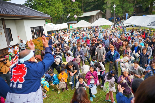 大勢の人々が屋外の広場に集まり、法被姿の人が豆や菓子を撒き、観客が手を伸ばして受け取ろうとしている地域のお祭りの様子を写した写真