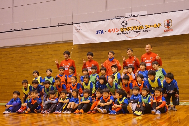 体育館の中央で赤いユニフォームを着たコーチたちと、色分けされたビブスを着た大勢の子どもたちが並んで集合写真を撮っている写真