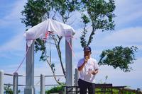 白い幕が掛けられた幸せの鐘の前で、男性がマイクを持ち話をしている除幕式の様子の写真