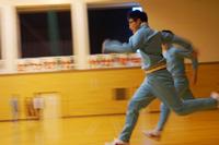 体育館の中でジャージを着た生徒が勢いよく全力で走っており、その後ろにも同じように走る生徒の姿が見える、運動中の一瞬をとらえた写真