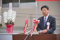 胸に白い胸章をつけた佐藤町長が、赤と白色のリボンが飾られたマイクの前に立ち挨拶をしている様子の写真