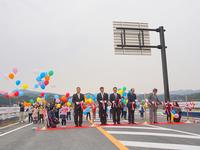 スーツ姿の男性たちが新しい道路の開通式で赤いリボンを持って並び、その後ろでは子どもたちや大人たちがカラフルな風船を持ってお祝いしている様子を写した写真