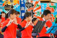 赤いシャツを着た吹奏楽部の学生たちが、カラフルな大漁旗の前で楽器を演奏している写真