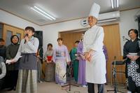 和服やエプロン姿の人々が室内で並び、マイクを持った女性が話す中、コック帽をかぶった高橋恒雄さんが前に立っている写真