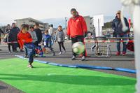 屋外で子どもがサッカーボールを蹴り、赤い服の大人が見守っているシュート体験の様子の写真