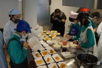 中学生たちがレストランで提供する料理を、台の上に並べられた食器に盛り付けている写真