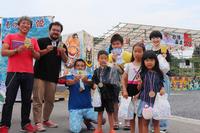 屋外で子どもたちと大人が笑顔で並び、手におもちゃや賞品を持って記念撮影している写真