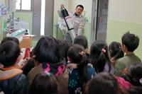 室内で消防署員が消火器の様な物を両手で持ち子どもたちに使い方を教えている写真