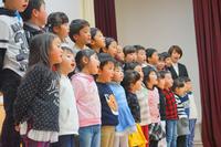 ステージの上に複数の子供たちが横2列に並び、観客の方を向いて立ち歌っている様子が写されている写真