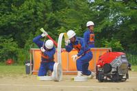 消防用ホースを取り扱う3人の団員が腰を落とし、ポンプ車のそばで水を汲もうとしている写真