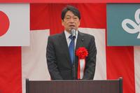 赤と白の幕の前で、赤いリボンを胸につけたスーツ姿の男性がマイクの前で式典の挨拶をしている写真