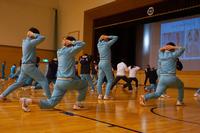 体育館のステージ前でジャージを着た生徒たちが手を頭の後ろで組みながら大きく足を踏み出し、姿勢を低くする運動を一斉に行っており、前方のスクリーンにはフォームの見本が映し出されているトレーニング中の様子を写した写真