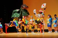 舞台の上で獅子舞の衣装を着た人物と扇子を持った踊り手が跳ねるように踊り、後ろでは法被姿の子どもたちが笛や太鼓で演奏している活気あふれる様子の写真