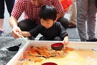 小さな男の子が大人と一緒にすくい網を水槽に入れて金魚すくいをしている写真