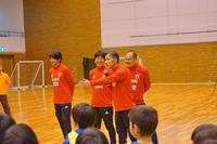 体育館で赤い服を着た4人の大人が前に立ち、子どもたちに向けて話している様子の写真