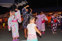 色とりどりの浴衣を着た女の子たちが両手を頭の上に上げて踊っている、夕暮れ時の盆踊りの写真