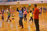 体育館で色とりどりのビブスを着た子どもたちがサッカーの練習をしており、大人が見守る中でボールを上に投げている場面の写真