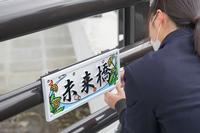 タンポポの絵がデザインされた「未来橋」と書かれた橋名板を、橋に設置している女子高校生の後ろ姿の写真