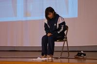 ステージ上で女性の方がパイプ椅子に座り、床に置いたタオルを足指を使って手繰り寄せる運動をしている写真