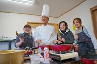 コック帽をかぶった高橋恒雄さんとエプロン姿の女性たちが調理器具を手に持ち、楽しそうに並んで料理をしている写真