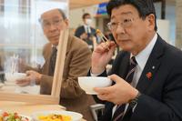 町長が白い器を左手に持ち、割りばしで持った食品を見ている写真