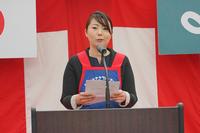 赤と白の幕の前で、エプロンを身に付けた女性がマイクの前に立ち、手元の原稿を読み上げている写真