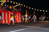 赤と白のやぐらの前で、若者たちが元気に飛び跳ねて現代風のダンスを踊っている夜のイベント写真