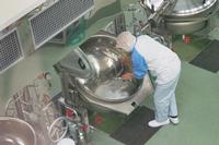 衛生帽子と作業着を着た給食調理員が、大きな業務用の回転釜をかがみ込んで丁寧に洗っている様子を上から撮影した、給食センター内での作業風景の写真