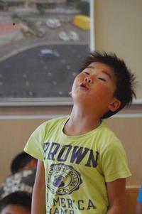 黄色いシャツを着た男の子が、顔を上に向けながら口をとがらせスイカの種を飛ばしている瞬間の写真