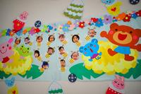 壁に貼られたカラフルな装飾が施されたボードに、動物のイラストや花の飾りとともに複数の子供たちの顔写真が整然と並べられている写真