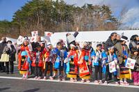 晴れた屋外で、色鮮やかな伝統衣装や法被着た子どもたちが並び、手に小旗を持って笑顔で立っている写真