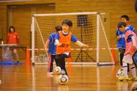 体育館で男の子がオレンジ色のビブスを着てサッカーボールを蹴ろうとしている瞬間の写真