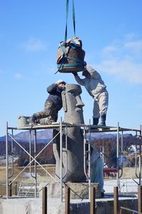 モアイ像の頭頂部にクレーンから吊り下げた円柱型の物が下ろされ、作業員が足場の上で手を伸ばしている様子を写した写真